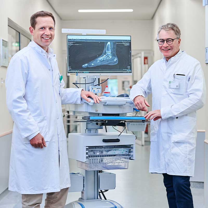 Zwei Ärzte in weißen Kitteln lächeln vor einem Monitor mit Röntgenbild eines Fußes im Krankenhaus.