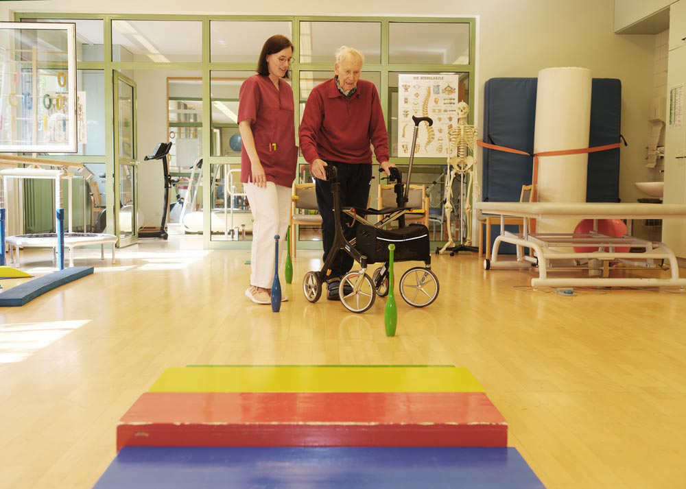 Ein älterer Mann mit Rollator übt das Gehen mit Hilfe einer Pflegerin in einem Rehabilitationsraum.
