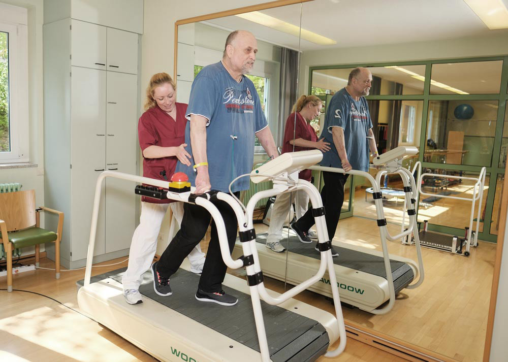 Ein Mann macht unter Anleitung einer Frau auf einem Laufband physiotherapeutische Übungen in einem hellen Raum.