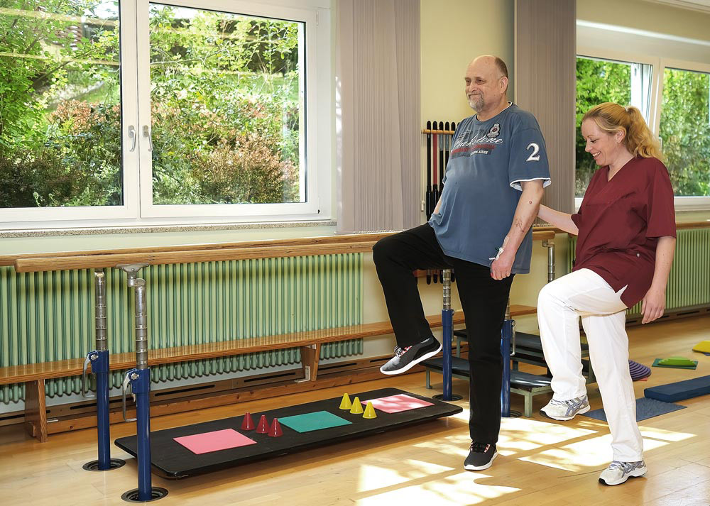 Ein Mann und eine Frau machen gemeinsam Reha-Übungen in einem Fitnessraum mit Fenstern und Übungsgeräten.