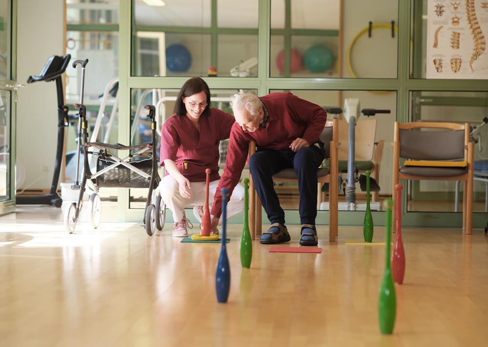 Ein älterer Mann macht Physiotherapie mit einer Betreuerin in einem Rehabilitationsraum.