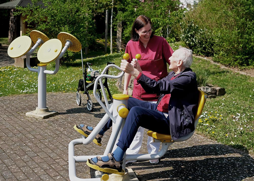 Eine Person im Rollstuhl unterstützt eine ältere Person auf einem Outdoor-Fitnessgerät in einem Park.