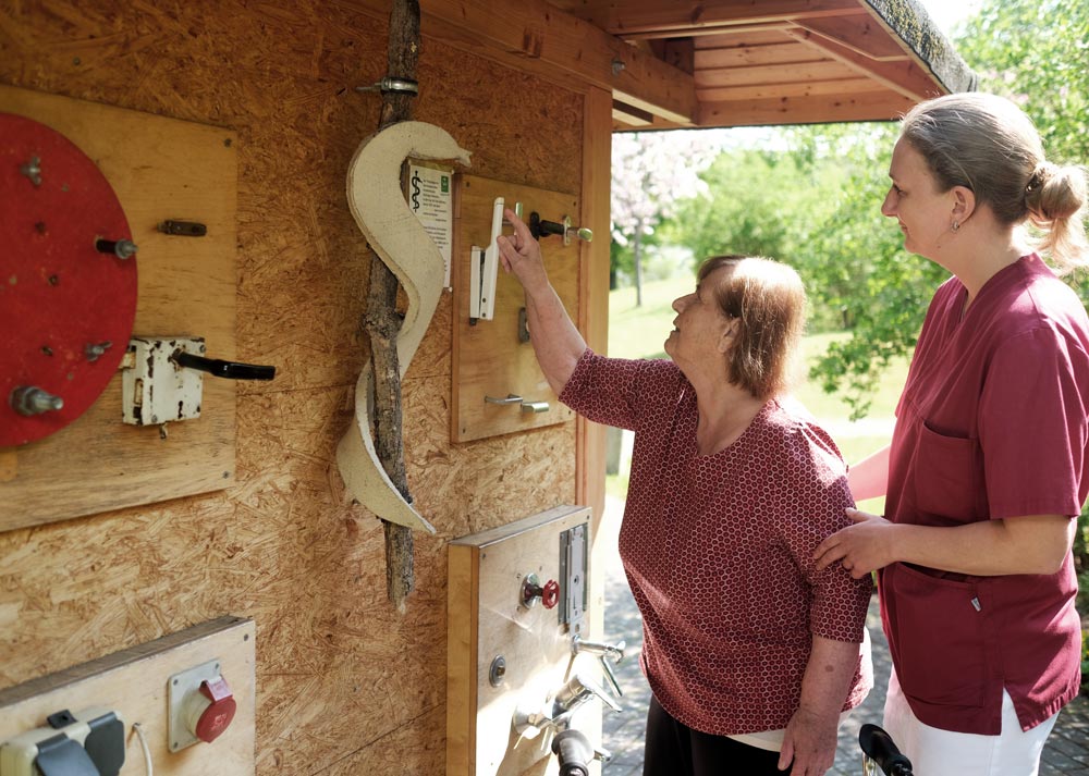 Eine Frau zeigt einer älteren Dame eine Holztafel mit Griffen in einem Garten. Sie stehen vor einer Wand mit mehreren Geräten.