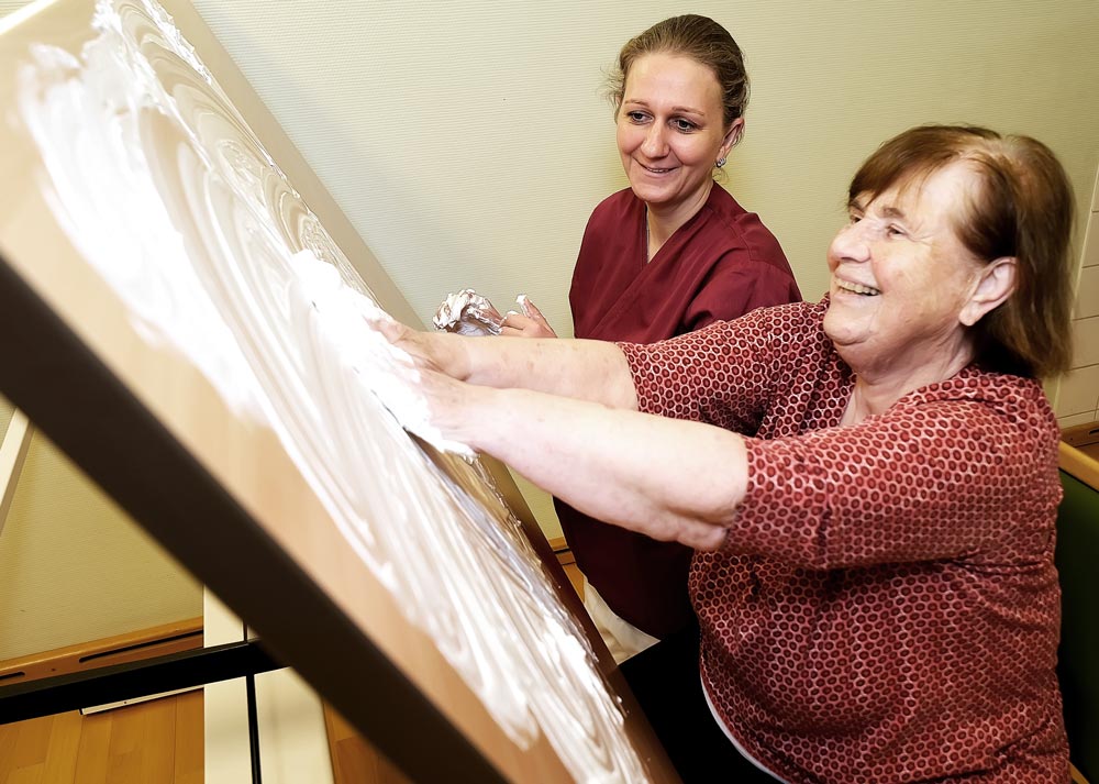 Eine ältere Frau malt mit weißer Farbe auf einer Leinwand, begleitet von einer jüngeren Frau, die ihr hilft und lächelt.