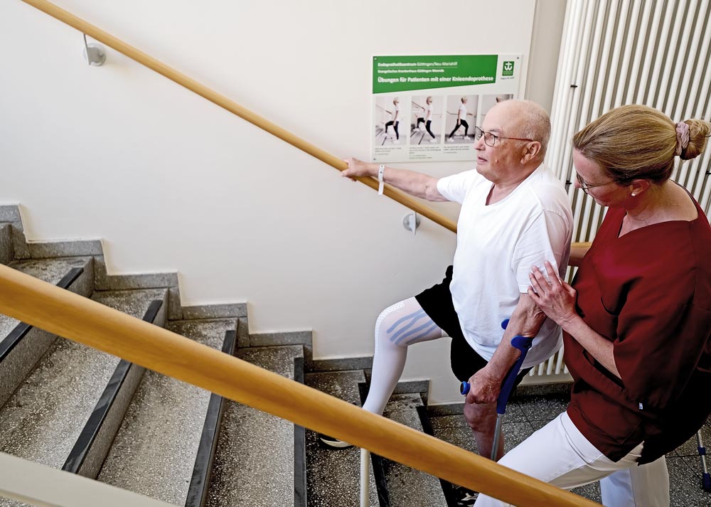 Ein Mann mit Beinprothese steigt eine Treppe hinauf, unterstützt von einer Frau. Poster mit Übungen an der Wand.