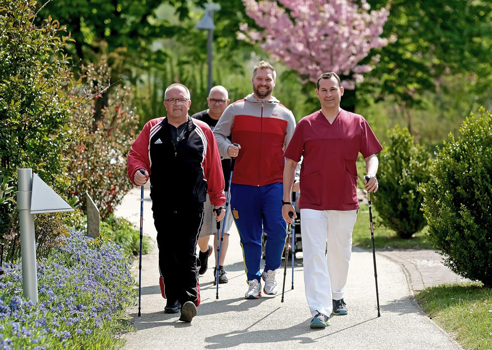 Eine Gruppe von Männern beim Nordic Walking auf einem Weg im Freien, umgeben von Bäumen und Blumen.