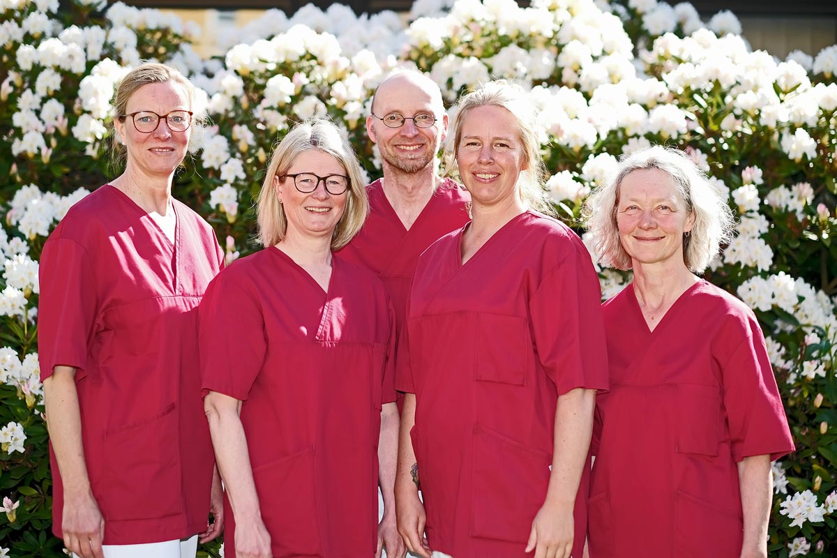 . Fünf Personen in roten Kitteln lächeln vor blühenden Büschen.
