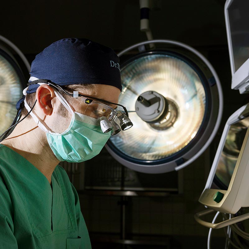 Ein Chirurg im Operationssaal mit Mundschutz, Kittel und Lupenbrille vor OP-Lampen.