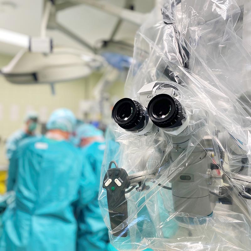 Ein Operationssaal mit Chirurgen in grüner Kleidung im Hintergrund und einem chirurgischen Mikroskop im Vordergrund.
