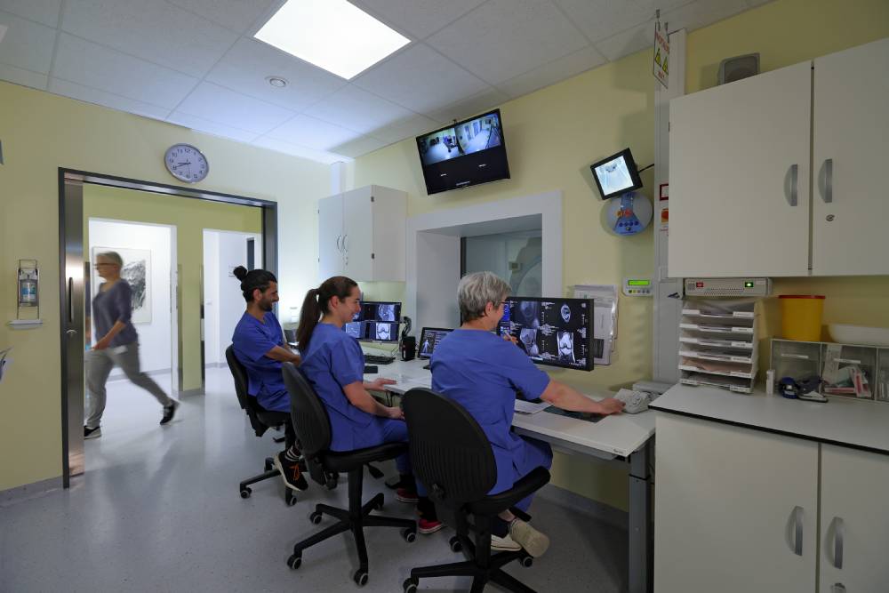 Ärzte in einem medizinischen Kontrollraum mit Monitoren, einer Uhr an der Wand und jemandem, der durch den Raum geht.