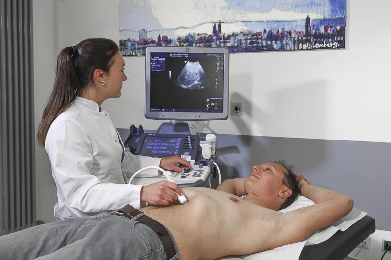 Eine Ärztin führt eine Ultraschalluntersuchung bei einem Patienten durch, der auf einer Liege liegt.