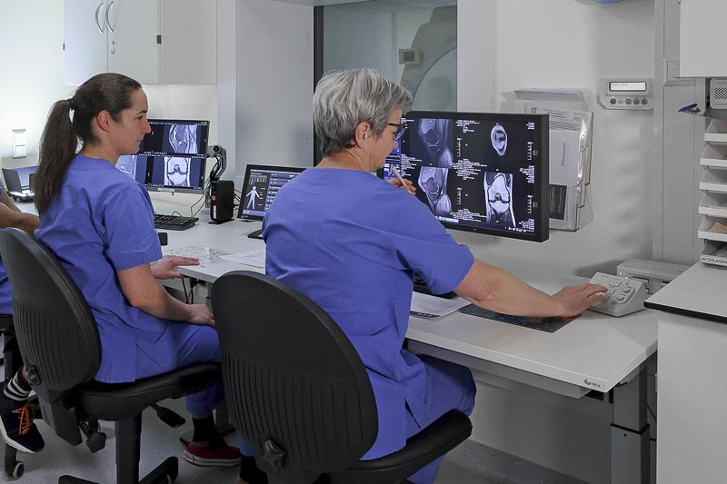 Zwei Personen in medizinischer Kleidung betrachten MRT-Bilder an Computermonitoren in einem Kontrollraum.