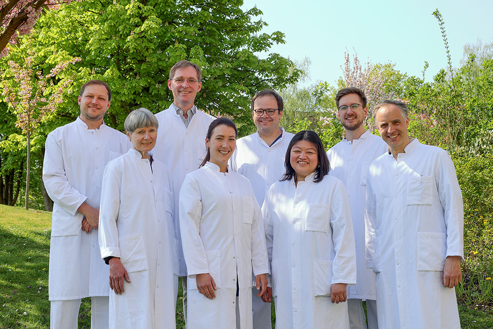 Eine Gruppe von Menschen in weißen Kitteln lächelt im Freien, umgeben von Bäumen.