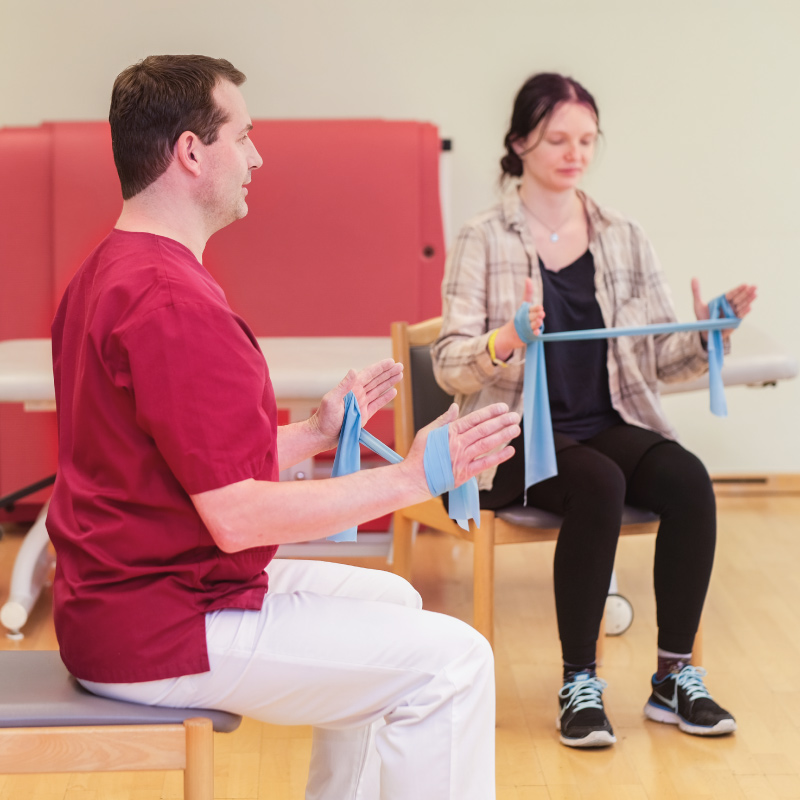 Zwei Menschen machen Physiotherapie-Übungen mit einem blauen Widerstandsband auf Stühlen im Raum.