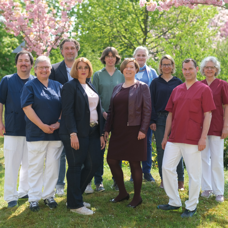 Eine Gruppe von Personen steht draußen vor blühenden Bäumen, lächelt, und trägt unterschiedliche Kleidung.