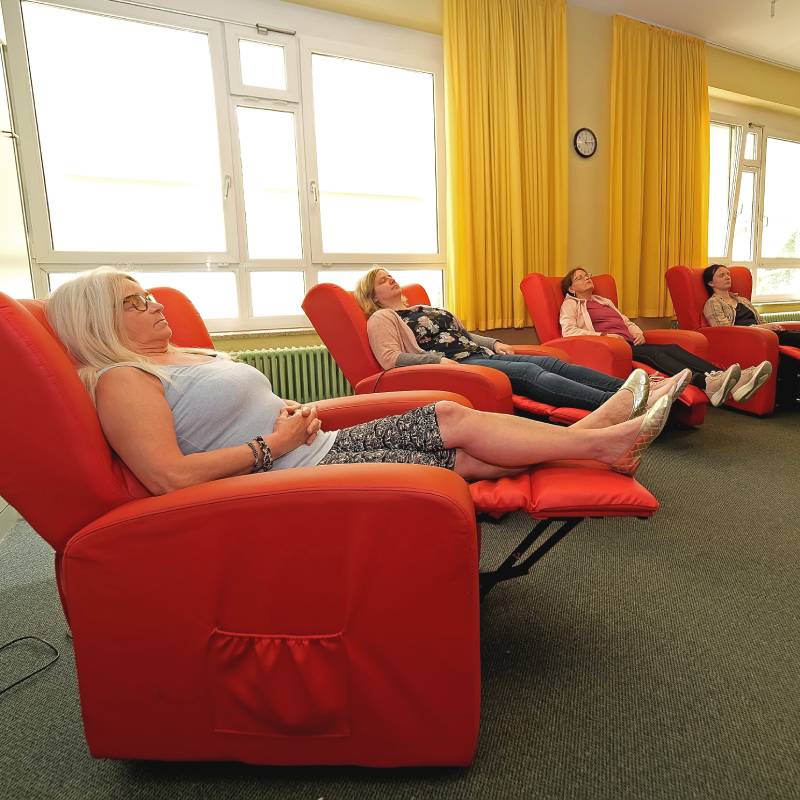 Vier Personen entspannen in roten Liegesesseln in einem Raum mit Fenstern und gelben Vorhängen.