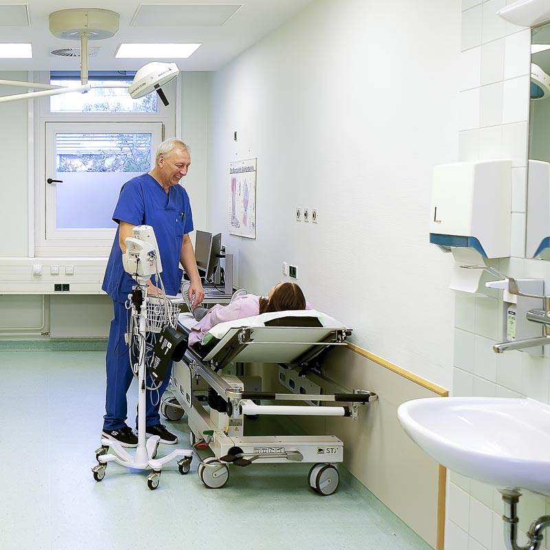 Ein Arzt in blauer Kleidung kümmert sich um einen Patienten auf einer Trage in einem Krankenhauszimmer.