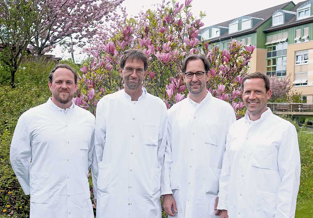 Vier Männer in weißen Kitteln stehen vor einem blühenden Baum im Freien.