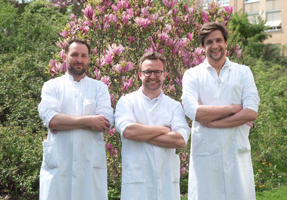 Drei Männer in weißen Kitteln stehen armverschränkt vor blühendem Baum im Freien.