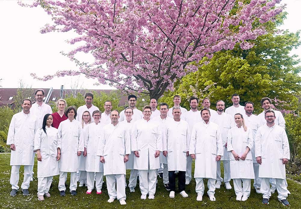 Eine Gruppe von Personen in weißen Kitteln steht vor einem blühenden Baum im Freien.