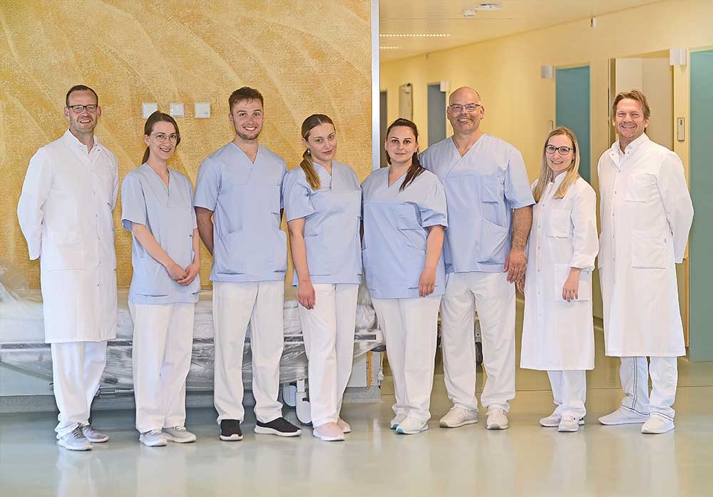 Gruppe von acht medizinischen Fachleuten in blauer und weißer Arbeitskleidung steht in einem Flur.