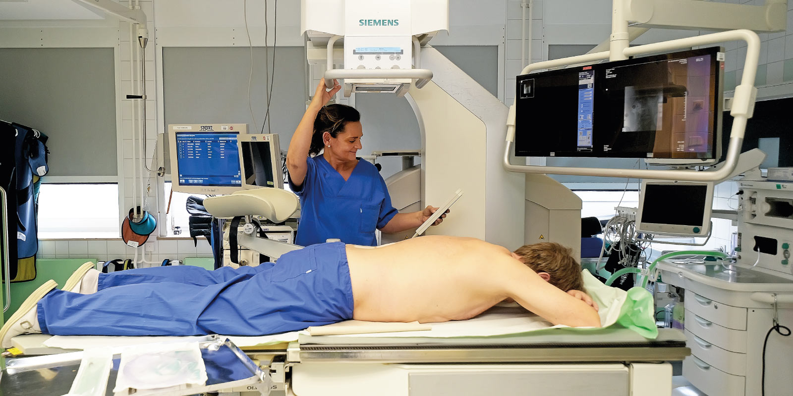 Ein Patient liegt auf einem Tisch in einem medizinischen Röntgenraum, ein Arzt in blauer Kleidung bedient Geräte.