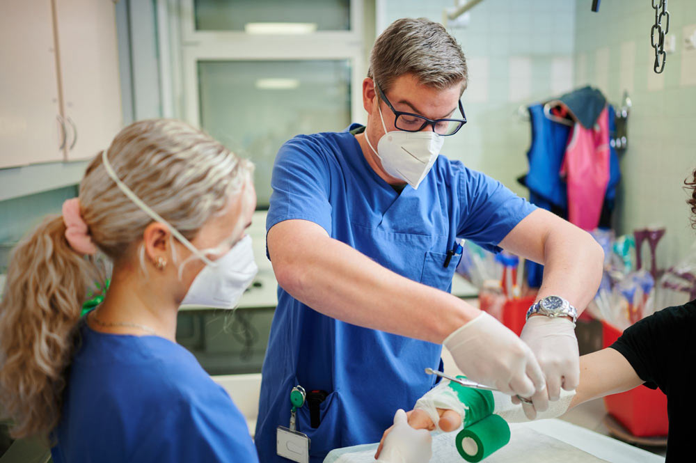 Ein Arzt legt einem Patienten einen grünen Verband an, während eine Assistentin zuschaut. Beide tragen Masken.