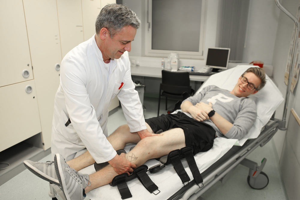 Ein Arzt untersucht das Bein eines lächelnden Patienten auf einer Krankenliege.