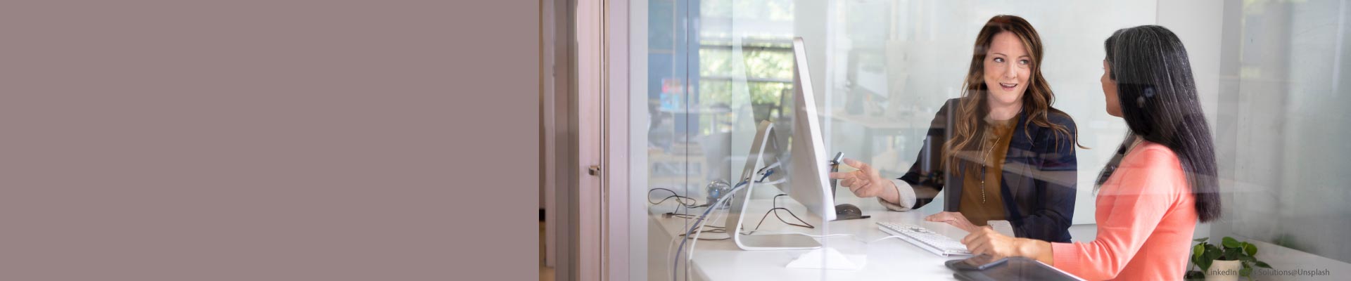 Zwei Frauen sitzen an einem Schreibtisch in einem Büro und unterhalten sich; sie arbeiten am Computer.