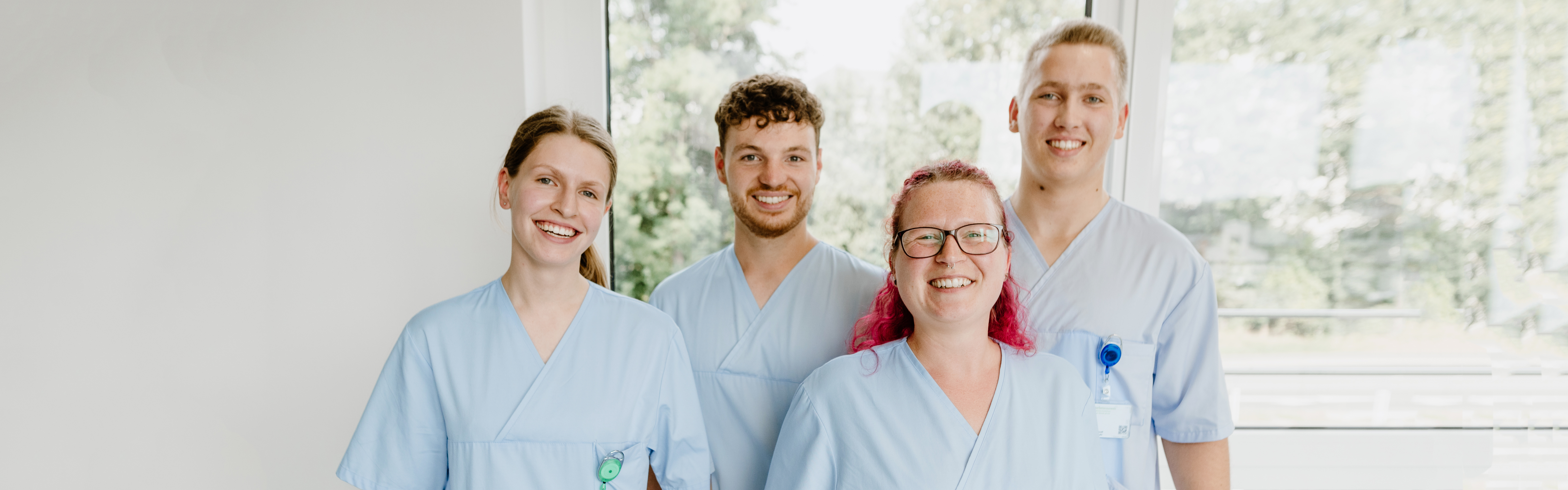 Eine Gruppe von vier Personen in blauer medizinischer Kleidung lächelt vor einem Fenster.