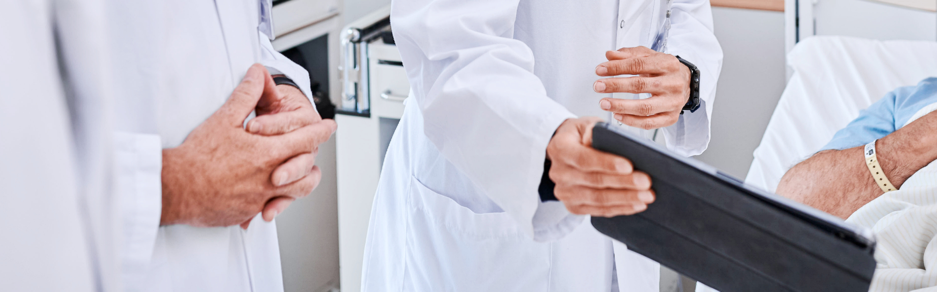 Ein Arzt mit Tablet spricht mit einem Patienten im Bett. Hände und weiße Kittel sind sichtbar.