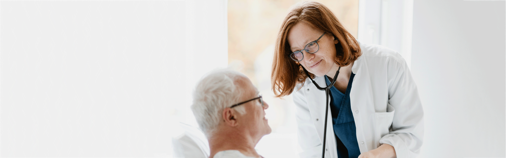 Eine Ärztin mit Stethoskop untersucht einen älteren Patienten und lächelt dabei freundlich.