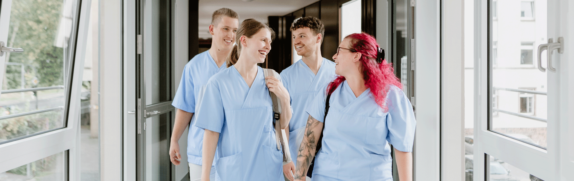 Vier Personen in blauen Krankenhausuniformen gehen lächelnd zusammen einen Gang entlang.