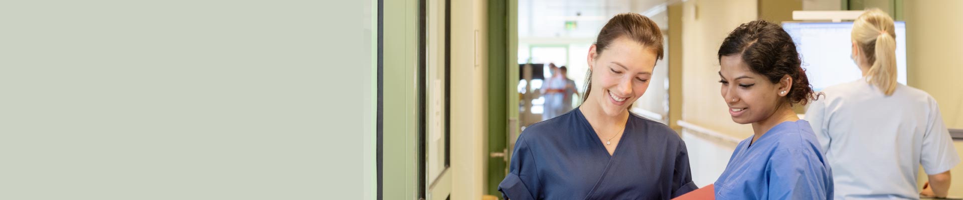 Zwei Personen in Krankenhauskleidung lächeln und betrachten ein Dokument; eine weitere Person arbeitet im Hintergrund.