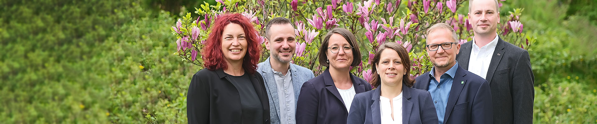 Eine Gruppe von sechs Personen steht lächelnd vor einem blühenden Strauch im Freien.
