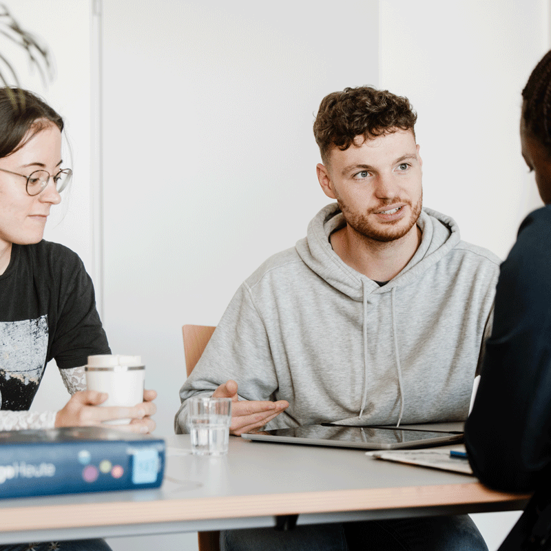 Drei Personen unterhalten sich an einem Tisch mit Getränken und Dokumenten.