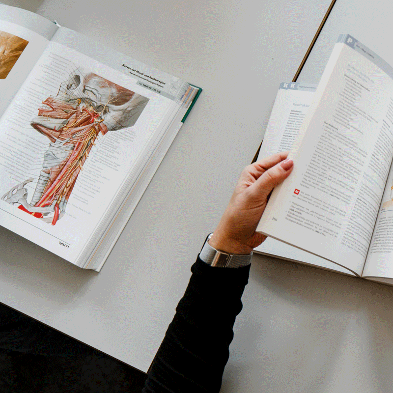 Eine Person blättert durch ein medizinisches Buch mit anatomischen Diagrammen auf einem Tisch.