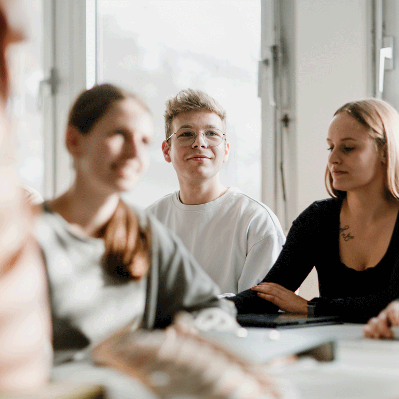 Drei Personen sitzen an einem Tisch in einem hellen Raum und hören aufmerksam zu.