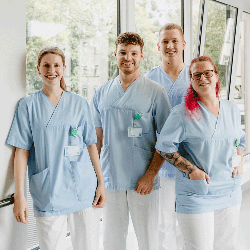 Vier lächelnde Menschen in Krankenhauskleidung stehen nebeneinander vor einem Fenster.