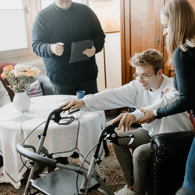 Eine Person hilft einer anderen, einen Rollator zu benutzen. Ein Dritter hält ein Tablet. Tisch im Hintergrund.