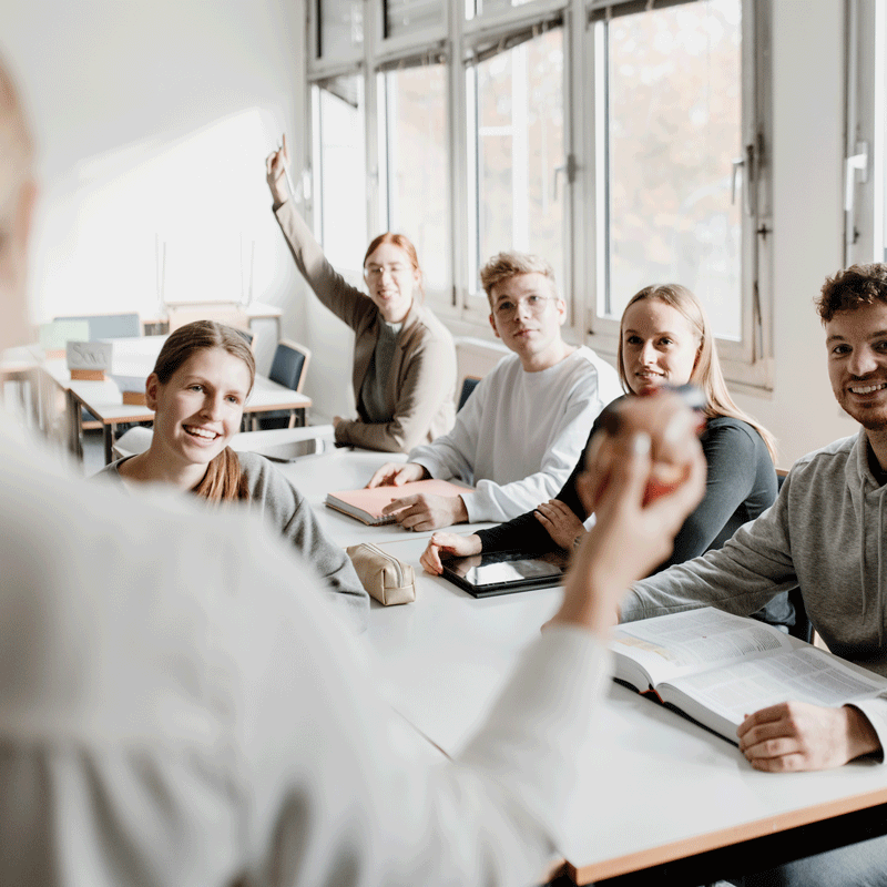 Eine Gruppe von Studenten sitzt in einem Klassenzimmer. Eine Person hebt die Hand, während ein Lehrender spricht.