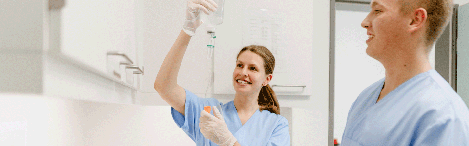 Zwei Personen in blauen Kitteln; eine hält eine Infusionsflasche und beide lächeln.
