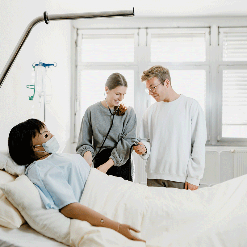 Medizinstudenten üben an einer Patientensimulationspuppe im Krankenhauszimmer; Frau und Mann mit Stethoskop.