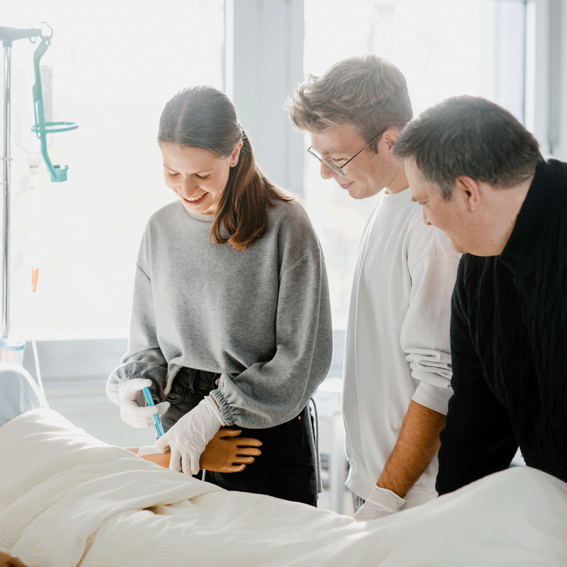 Drei Personen in einem Krankenhauszimmer, eine führt eine medizinische Tätigkeit bei einem Patienten aus.