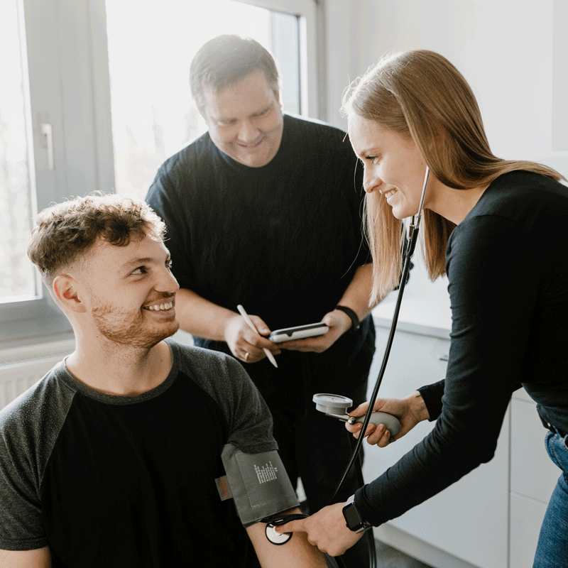 Eine Ärztin misst den Blutdruck eines lächelnden Mannes, während ein weiterer Mann Notizen macht.