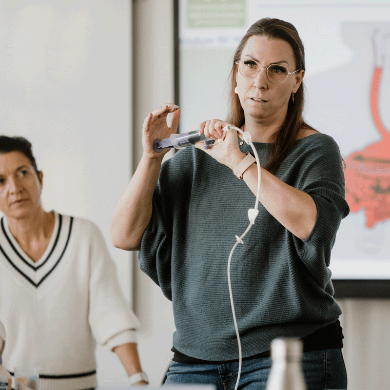 Zwei Frauen in einem Seminarraum, eine hält eine Spritze mit einem Schlauch, die andere schaut zu.