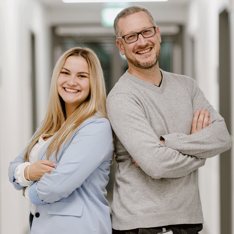 Zwei lächelnde Personen stehen Rücken an Rücken in einem Flur, beide mit verschränkten Armen.