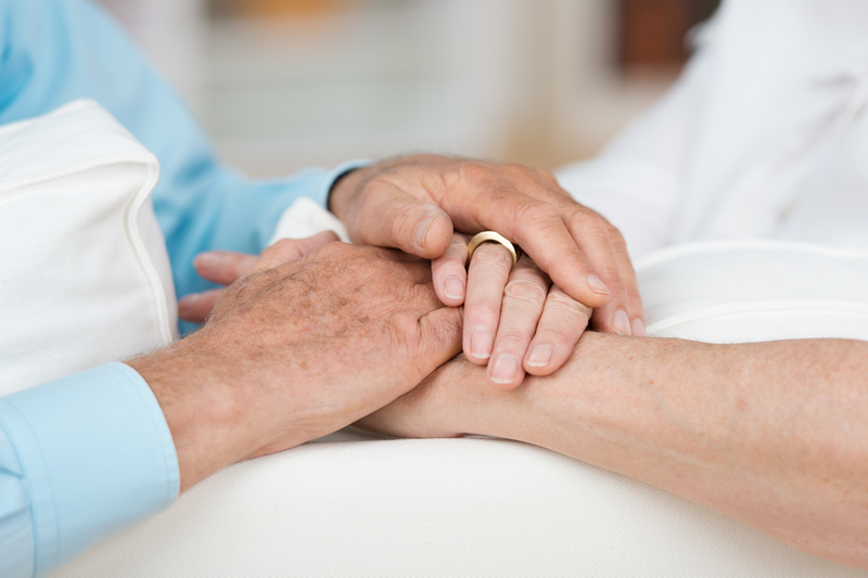 Zwei ältere Menschen halten sich zärtlich die Hände, eine trägt einen Ehering.