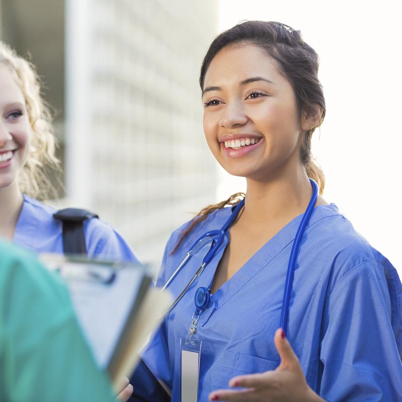 Eine lächelnde Medizinstudentin in blauer Kleidung mit Stethoskop spricht mit Kollegen.
