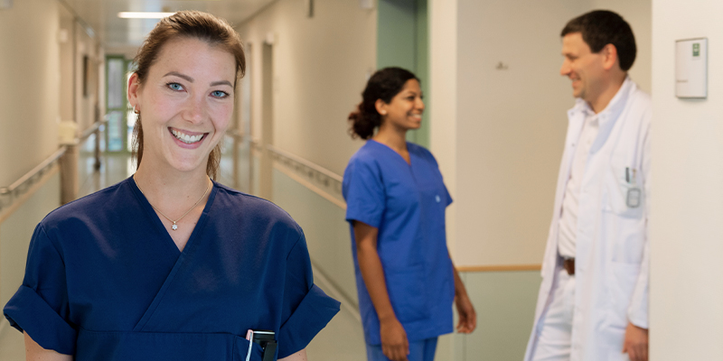 Drei Medizinfachkräfte in einem Krankenhausflur, freundlich lächelnd, zwei im Gespräch im Hintergrund.
